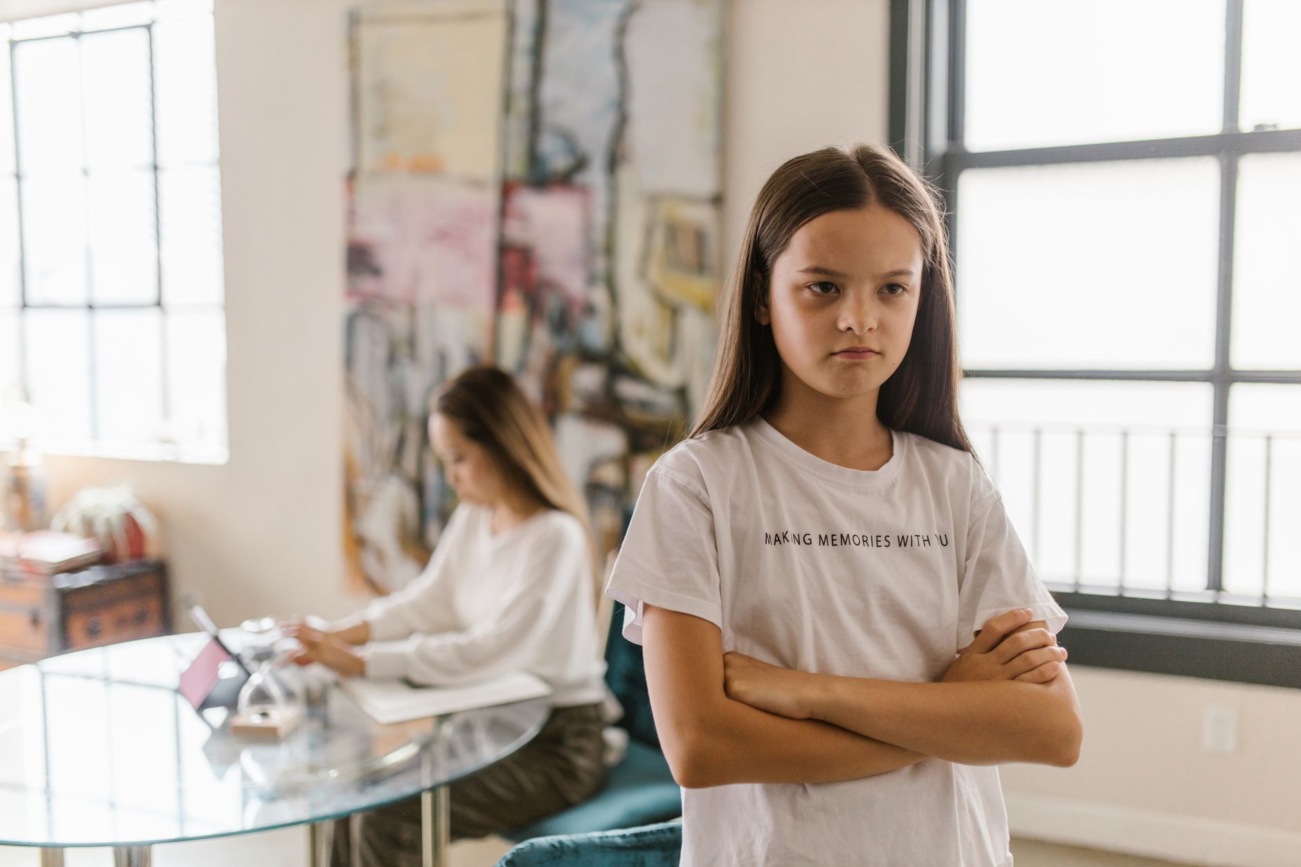 A young girl standing with her arms crossed while a parent works in the background